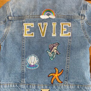 May include: A light blue denim jacket with a rainbow, clouds, and the name "EVIE" in gold letters. The jacket also has patches of a mermaid, a seashell, and a starfish.