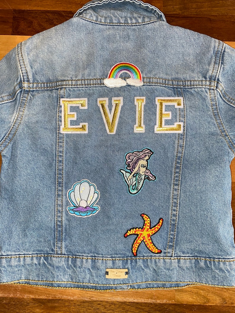 May include: A light blue denim jacket with a rainbow, clouds, and the name "EVIE" in gold letters. The jacket also has patches of a mermaid, a seashell, and a starfish.