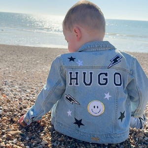 Peut inclure: Une veste en jean bleu clair avec des patchs noirs et blancs, dont un éclair, des étoiles et un smiley. La veste porte le nom "HUGO" en lettres noires au dos.