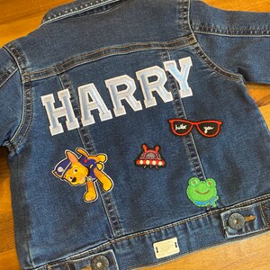 May include: A blue denim jacket with the name "HARRY" in white letters on the back. The jacket has several patches, including a cartoon dog, a spaceship, a frog, and a pair of sunglasses.