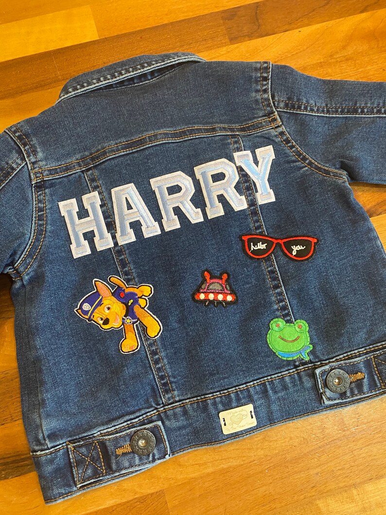 May include: A blue denim jacket with the name "HARRY" in white letters on the back. The jacket has several patches, including a cartoon dog, a spaceship, a frog, and a pair of sunglasses.