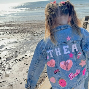 May include: A light blue denim jacket with the name "THEA" in white letters on the back. The jacket is decorated with various colorful patches, including a pink heart with sequins, a pink star, a pink smiley face, a pink car, a pink drink, and a pink Barbie logo.