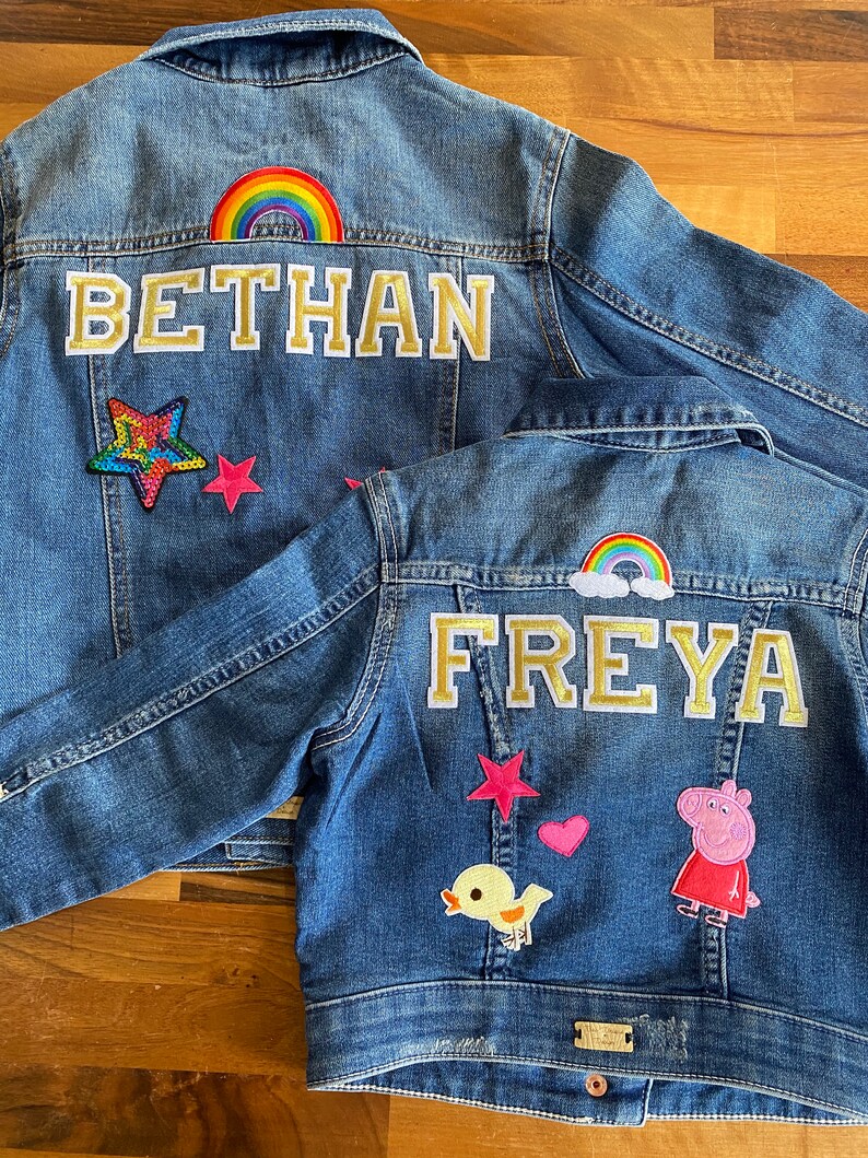 May include: Two denim jackets with colorful patches and embroidered names. The larger jacket has a rainbow, stars, and the name "BETHAN" embroidered in gold. The smaller jacket has a rainbow, a pink star, a pink heart, a yellow chick, a Peppa Pig patch, and the name "FREYA" embroidered in gold.