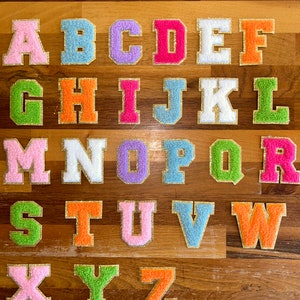 May include: A set of 26 colourful felt letters with gold glitter trim. The letters are arranged in alphabetical order on a wooden surface.