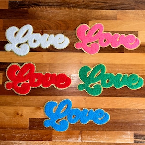 May include: Five fuzzy patches with the word "Love" in a retro font. The patches are in white, pink, red, green, and blue with gold glitter outlining the letters.