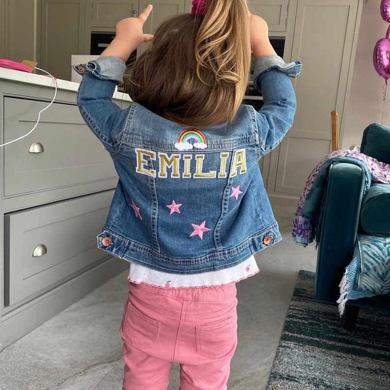 Op de afbeelding: Een blauw denim jack met een regenboog en de naam "EMILIA" geborduurd op de rug. Het jack heeft roze stervormige patches.