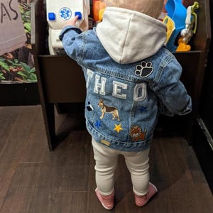 Puede incluir: Un niño pequeño lleva una chaqueta vaquera azul con el nombre "THEO" bordado en la espalda. La chaqueta tiene varios parches, incluyendo una huella de pata, un perro y una estrella. El niño lleva pantalones blancos y calcetines a rayas rojas y blancas.