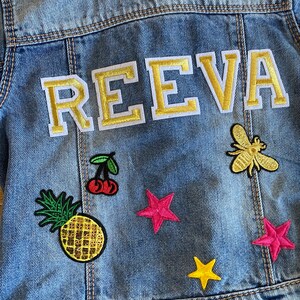 May include: A light blue denim jacket with a gold embroidered name "REEVA" on the back. The jacket has various colorful patches including a pineapple, cherries, stars, and a bee.