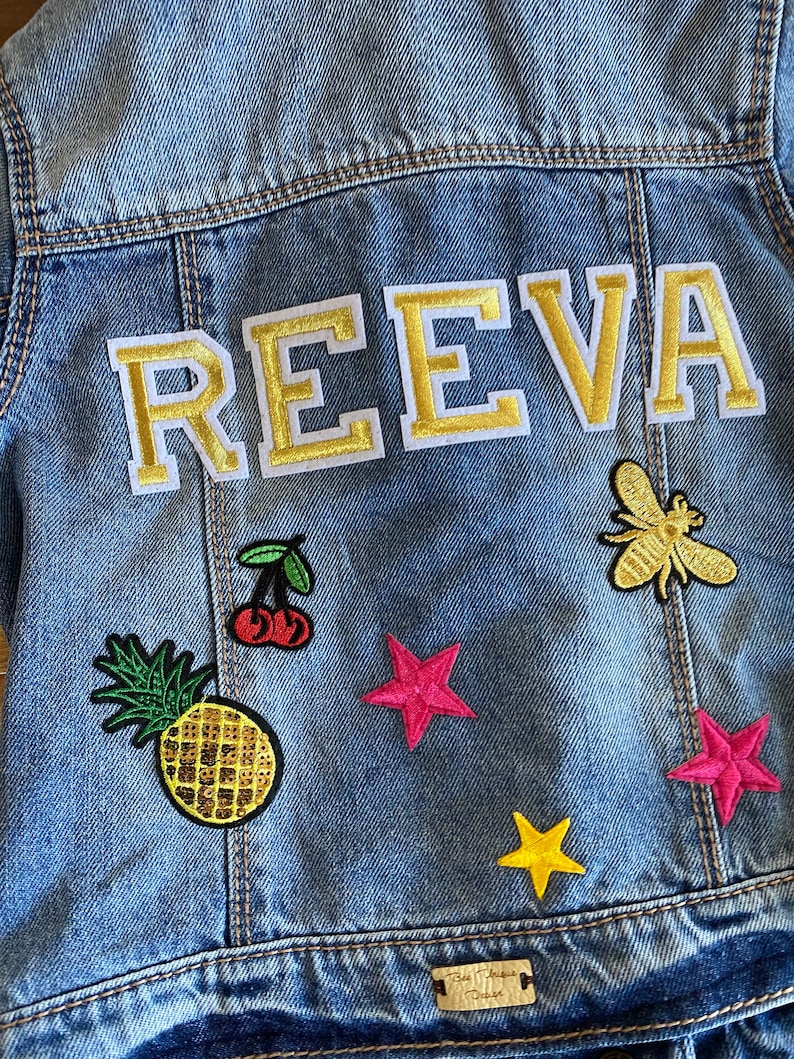 May include: A light blue denim jacket with a gold embroidered name "REEVA" on the back. The jacket has various colorful patches including a pineapple, cherries, stars, and a bee.