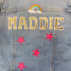 Op de afbeelding: Een lichtblauw denim jack met een regenboog- en wolkpatch, een gouden "MADDIE"-patch en vier roze sterpatches.