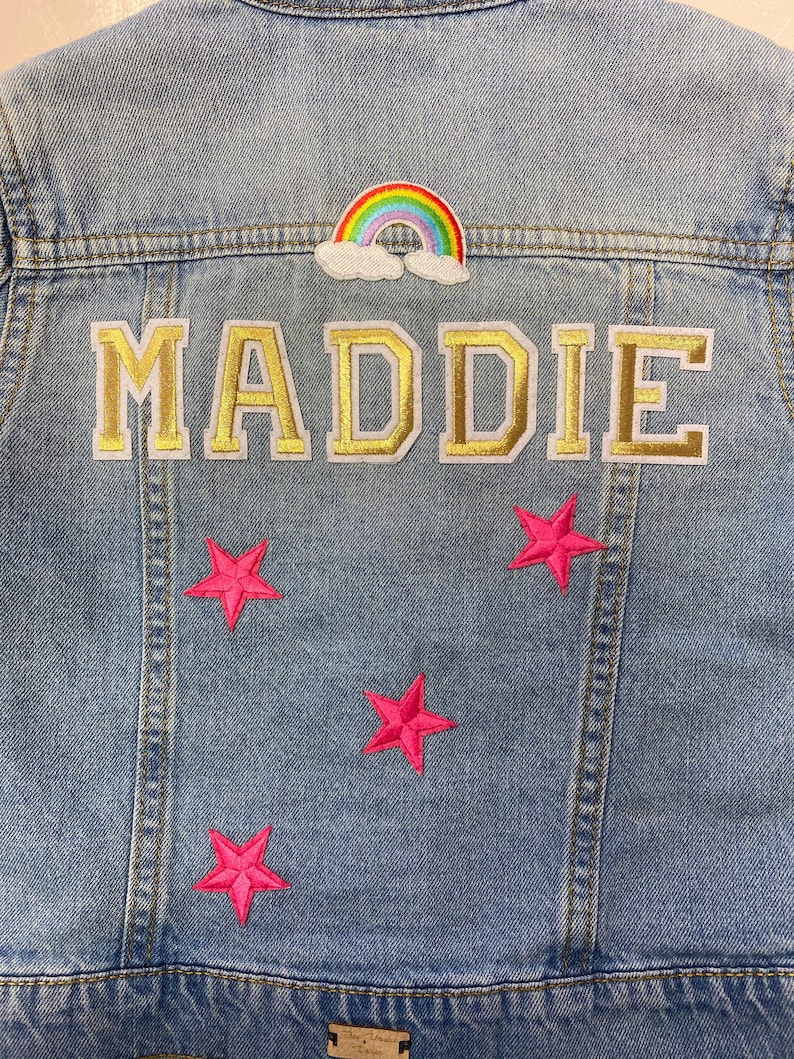Op de afbeelding: Een lichtblauw denim jack met een regenboog- en wolkpatch, een gouden "MADDIE"-patch en vier roze sterpatches.