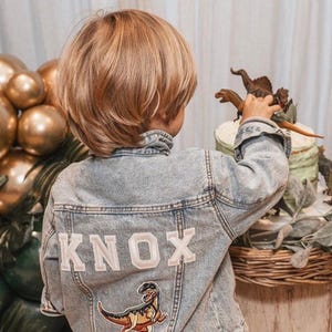 May include: A young boy wearing a light blue denim jacket with the name "KNOX" embroidered on the back. The jacket has a dinosaur patch on the lower back. The boy is reaching for a dinosaur cake topper.