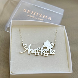 Peut inclure: Un collier en argent avec un pendentif Hello Kitty et le nom "Anastasia" écrit en cursive. Le collier est présenté dans une boîte blanche avec les mots "SEHISHA JEWELLERY" imprimés sur le couvercle.