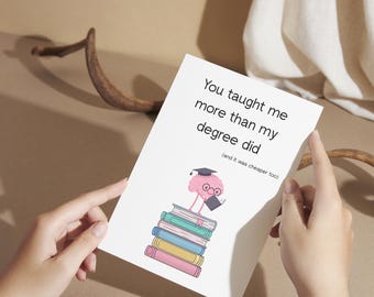 May include: A white greeting card held by hands, featuring the text "You taught me more than my degree did" and a cartoon flamingo wearing a graduation cap and glasses, standing on a stack of books.