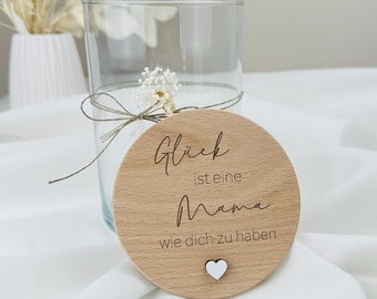 以下が含まれることがあります： 「Glück ist eine Mama wie dich zu haben」（幸せはあなたのようなお母さんを持つことです）と書かれた丸い木製の看板と小さな白いハート。