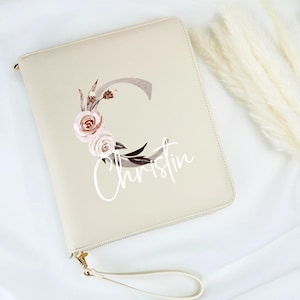 Könnte beinhalten: Eine beige Leder-Reißverschlusstasche mit einem weißen Blumendesign und dem Namen "Christin" darauf gedruckt. Die Tasche hat einen weißen Riemen.