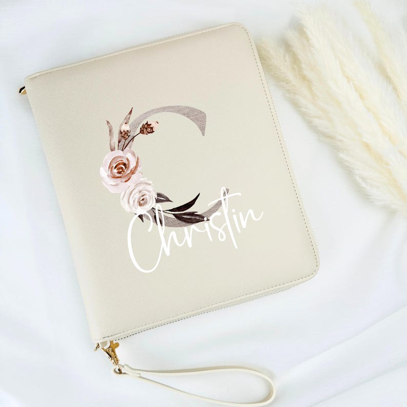 May include: A beige leather zipped pouch with a white floral design and the name "Christin" printed on it. The pouch has a white strap.