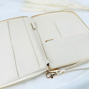 May include: A cream-coloured zippered pouch with multiple pockets and a gold zipper. The pouch has a gold hook and a cream-coloured strap.