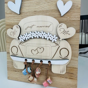 以下が含まれることがあります： 花、ハート、そして「just married」の文字で飾られた、白い車の切り抜きが施された木製の看板。車は後ろに小さな白いハートが2つ、側面に「Mr.」と「Mrs.」の文字が書かれている。車の底から茶色の紐が垂れ下がり、巻かれたお金が付けられている。