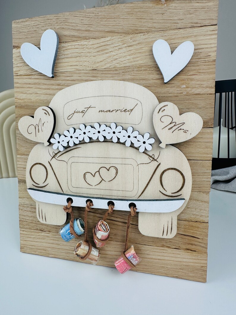 以下が含まれることがあります： 花、ハート、そして「just married」の文字で飾られた、白い車の切り抜きが施された木製の看板。車は後ろに小さな白いハートが2つ、側面に「Mr.」と「Mrs.」の文字が書かれている。車の底から茶色の紐が垂れ下がり、巻かれたお金が付けられている。
