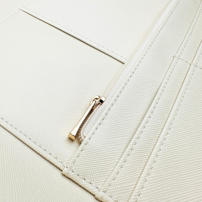 May include: Close-up of a white leather wallet with a gold zipper. The wallet has multiple compartments and is stitched with white thread.