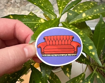 May include: A sticker featuring a cartoon illustration of a red couch with a purple background. The couch has a tufted back and armrests, and is set against a white and black base. The sticker is held in front of a green plant.