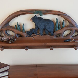 Puede incluir: Una estanter&iacute;a de pared de madera con un dise&ntilde;o tallado de oso y cachorro. La estanter&iacute;a tiene cuatro ganchos y est&aacute; hecha de madera marr&oacute;n oscura.