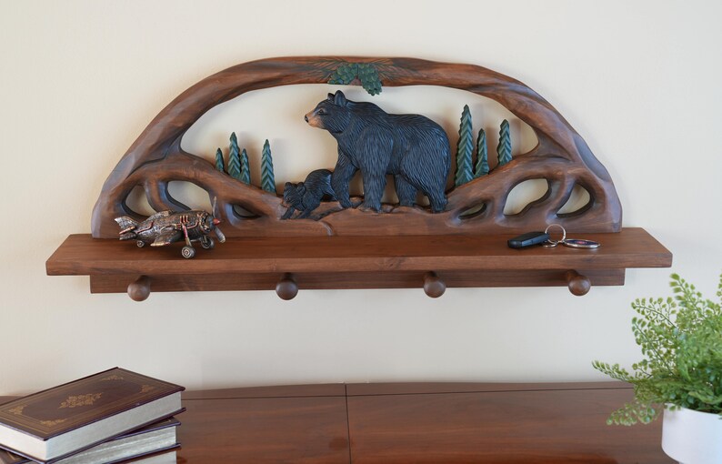 Puede incluir: Una estanter&iacute;a de pared de madera con un dise&ntilde;o tallado de oso y cachorro. La estanter&iacute;a tiene cuatro ganchos y est&aacute; hecha de madera marr&oacute;n oscura.