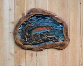 May include: A carved wooden wall hanging depicting a trout jumping out of the water. The trout is painted with realistic colors, including red, orange, and blue. The background is a blue and green water scene with rocks and a log.