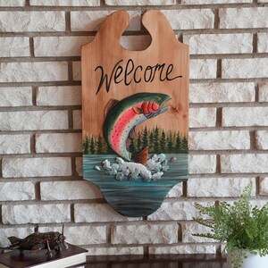 May include: Wooden welcome sign with a painted rainbow trout jumping out of the water. The sign has the word "Welcome" written in cursive on the top.