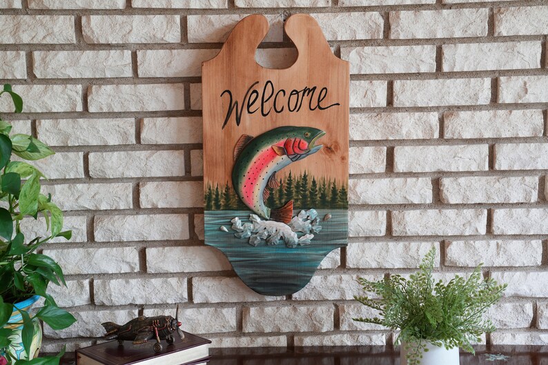 May include: Wooden welcome sign with a painted rainbow trout jumping out of the water. The sign has the word "Welcome" written in cursive on the top.