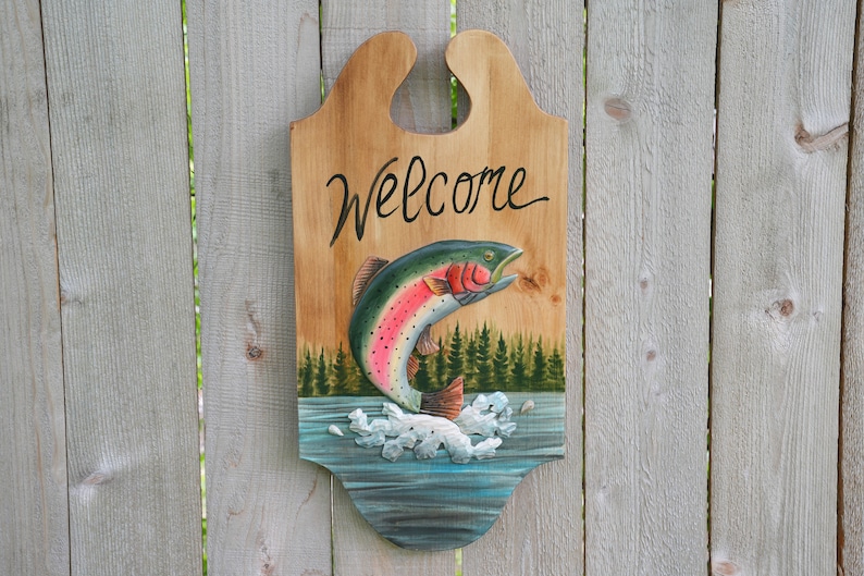 May include: Wooden welcome sign with a carved rainbow trout jumping out of the water. The sign is painted with a blue water background, green trees, and white foam. The words "Welcome" are written in black cursive on the sign.
