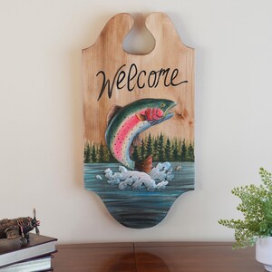 May include: Wooden welcome sign with a painted rainbow trout jumping out of the water. The sign has a cutout for hanging and says "Welcome" in black cursive lettering.