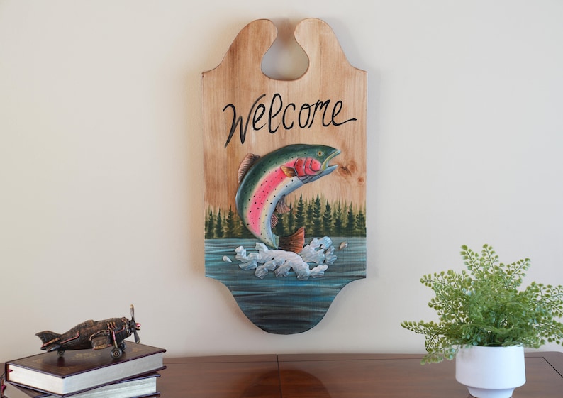 May include: Wooden welcome sign with a painted rainbow trout jumping out of the water. The sign has a cutout for hanging and says "Welcome" in black cursive lettering.