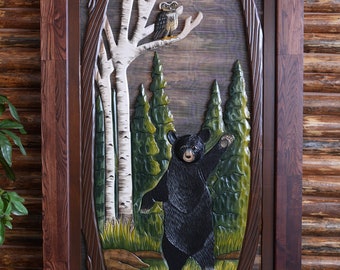 May include: A carved wooden door panel with a detailed scene of a black bear standing in a forest with birch trees and an owl perched on a branch. The panel is framed in dark wood.