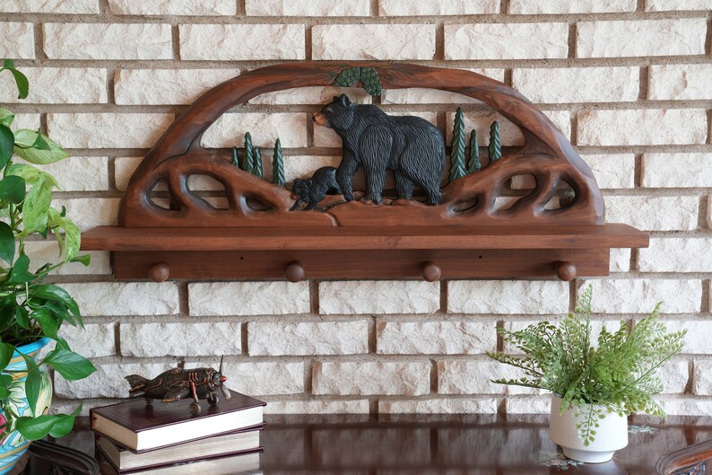 Puede incluir: Una estanter&iacute;a de madera con un dise&ntilde;o tallado de un oso y un cachorro. La estanter&iacute;a tiene dos ganchos en la parte inferior y est&aacute; montada en una pared de ladrillo.