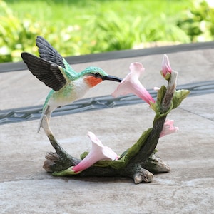 May include: A detailed figurine of a hummingbird with outstretched wings, poised to feed from pink trumpet-shaped flowers. The bird has green, white, and black plumage, with a red throat. The flowers are attached to a brown, branch-like base.