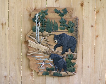 May include: A wooden wall carving depicting two black bears in a forest setting. The bears are carved in a realistic style and are shown standing near a stream. The carving is painted in shades of brown, green, and blue.
