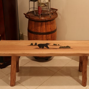 May include: A rustic wooden bench with a black bear and green trees carved into the top. The bench has two legs on each side.