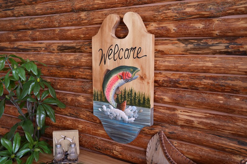 May include: Wooden welcome sign with a painted image of a rainbow trout jumping out of the water. The sign has a hole at the top for hanging and says "Welcome" in cursive.