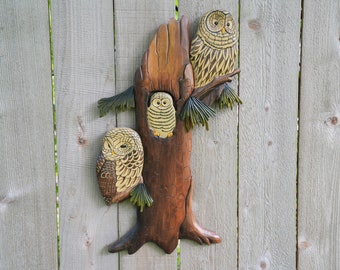 May include: A wooden carving of three owls perched on a tree trunk. The owls are painted in shades of brown, white, and yellow. The tree trunk is painted in shades of brown and has a textured surface.
