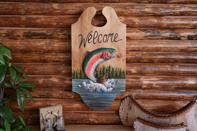 May include: Wooden welcome sign with a carved and painted rainbow trout jumping out of the water. The sign has a cutout handle at the top and says "Welcome" in cursive.