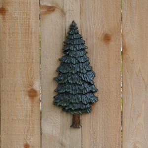May include: A dark green wooden pine tree sculpture mounted on a light brown wooden fence.