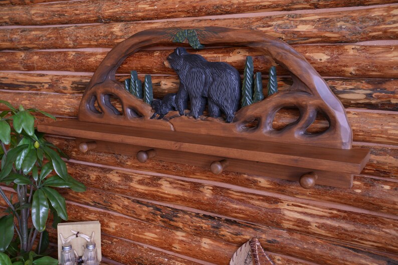 Puede incluir: Una estanter&iacute;a de madera con un dise&ntilde;o tallado de oso y pino. La estanter&iacute;a tiene tres ganchos y est&aacute; te&ntilde;ida de un color marr&oacute;n oscuro.
