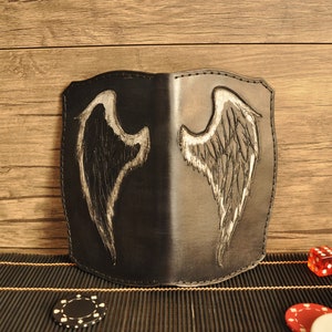 Puede incluir: Cartera de cuero negro con un diseño de alas de ángel pintado a mano. La cartera está abierta, revelando el interior. El fondo incluye una superficie de madera, dados y fichas de póquer, lo que sugiere un tema de juegos o apuestas.