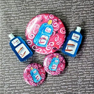 Könnte beinhalten: Eine Sammlung von blauen und roten Ansteckbuttons und einem Flaschenförmigen Pin. Die Pins zeigen ein blaues Flaschendesign mit dem Text "99,9%" und "DRAGONBREATH X". Der Hintergrund ist eine dunkelgraue Oberfläche mit handschriftlichem Text.