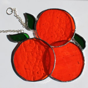May include: Three stained glass oranges with green leaves hanging from a silver chain. The oranges are a bright orange colour and the leaves are a dark green.