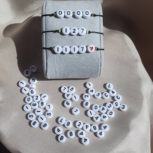 Puede incluir: Tres pulseras de cuerda negra con cuentas blancas que deletrean los números 0000, 127 y 1117 con una cuenta roja en forma de corazón al final de la tercera pulsera. También hay muchas cuentas blancas con números negros esparcidas sobre una superficie beige.