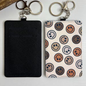 May include: Two rectangular keychains. One is black with a textured surface and slots. The other is off-white with a pattern of smiley faces in brown, orange, and black. Both have silver key rings.