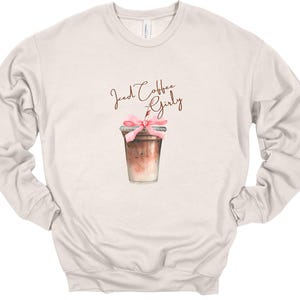 Puede incluir: Una sudadera beige con el texto "Iced Coffee Girly" en letras marrones. Una ilustraci&oacute;n de acuarela de una taza de caf&eacute; helado con una cinta rosa y una pajita est&aacute; centrada en la sudadera.